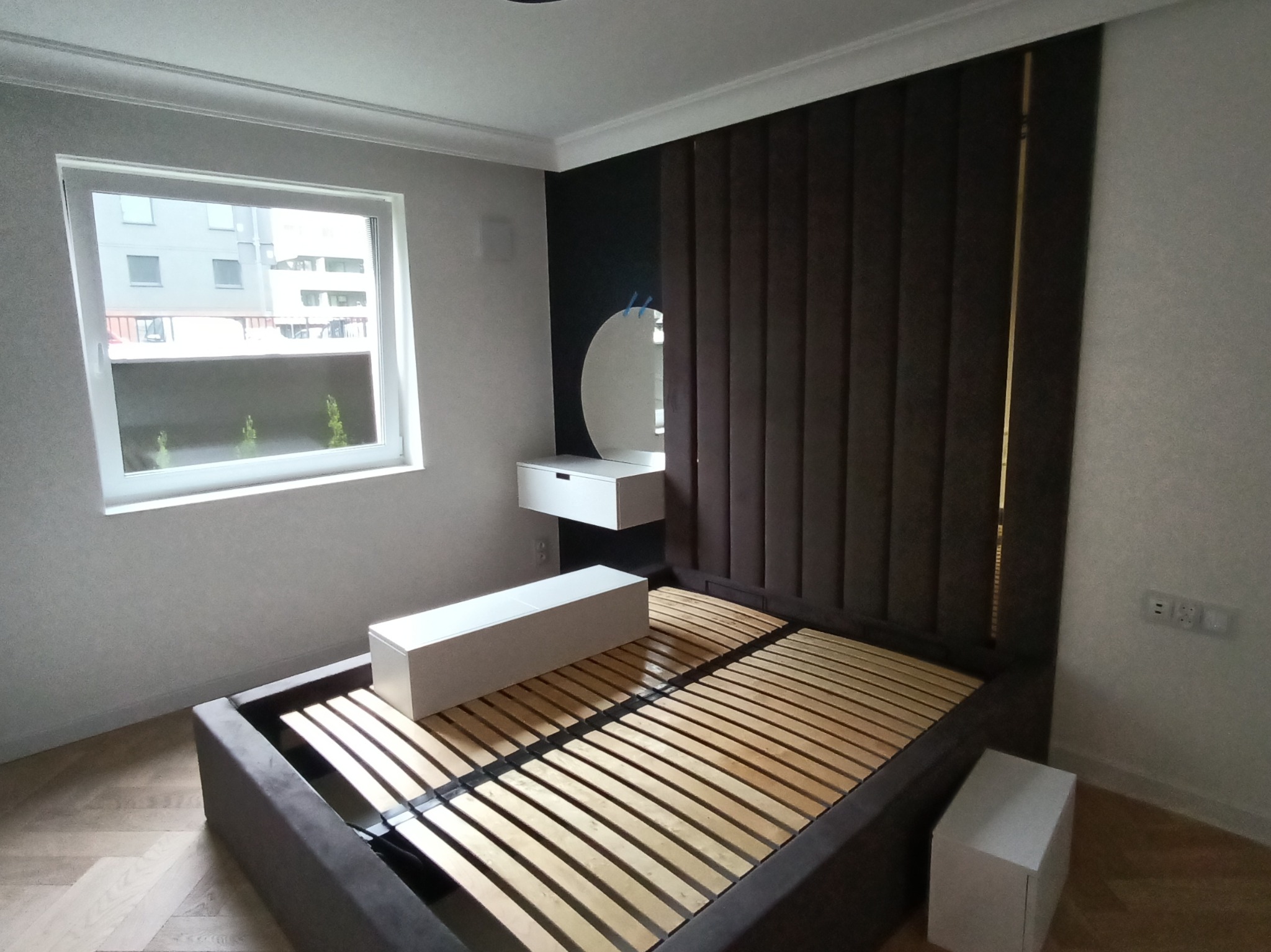 Nowoczesna sypialnia z łóżkiem z brązowym wezgłowiem, białą toaletką i lustrem. Parkiet w jodełkę. Widok na budynek przez okno. Minimalistyczny design.