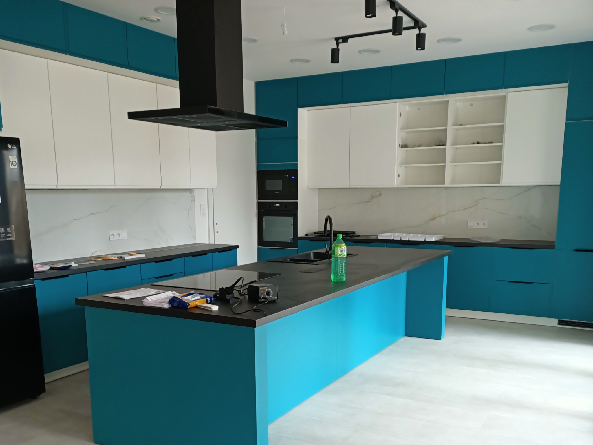 Nowoczesna kuchnia z turkusowymi i białymi szafkami, czarnym okapem i wyspą. Widoczne detale wykończenia i sprzęt AGD. Ujęcie z perspektywy wnętrza.