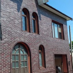 VOLODYMYR KRYSHTALSKYI KVEN - Dwupiętrowy dom z ciemnej cegły klinkierowej z łukowymi oknami, częściowo zasłonięty rusztowaniem. Widoczny fragment muru z cegły w tle i słup energetyczny.