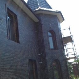 VOLODYMYR KRYSHTALSKYI KVEN - Elewacja z ciemnej cegły klinkierowej, widok z dołu na narożnik budynku z wieżyczką. Widoczne rusztowania i zieleń w tle. Charakterystyczne łukowe wnęki okienne.