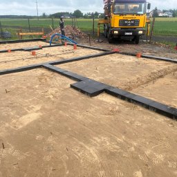 VOLODYMYR KRYSHTALSKYI KVEN - Przygotowany teren pod fundamenty z ułożonymi bloczkami, widoczne rury kanalizacyjne i zbrojenie. W tle betoniarka i pracownik budowlany.