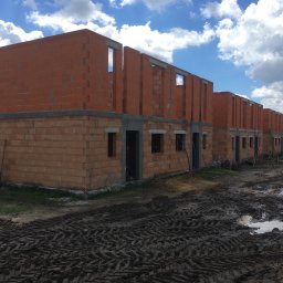 VOLODYMYR KRYSHTALSKYI KVEN - Rząd surowych, ceglanych budynków w trakcie budowy pod błękitnym niebem z chmurami. Widoczne ślady opon na błotnistym terenie.