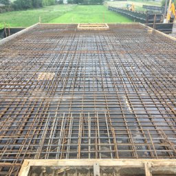 VOLODYMYR KRYSHTALSKYI KVEN - Zbrojenie płyty fundamentowej z widocznym deskowaniem i otoczeniem budowy. Widok z góry na siatkę z prętów zbrojeniowych, przygotowaną do zalania betonem.