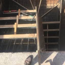 VOLODYMYR KRYSHTALSKYI KVEN - Zbrojenie fundamentów w szalunku z desek, widok z góry. Poziomica, młotek i buty robocze w kadrze. Elementy konstrukcyjne z betonu i cegły.