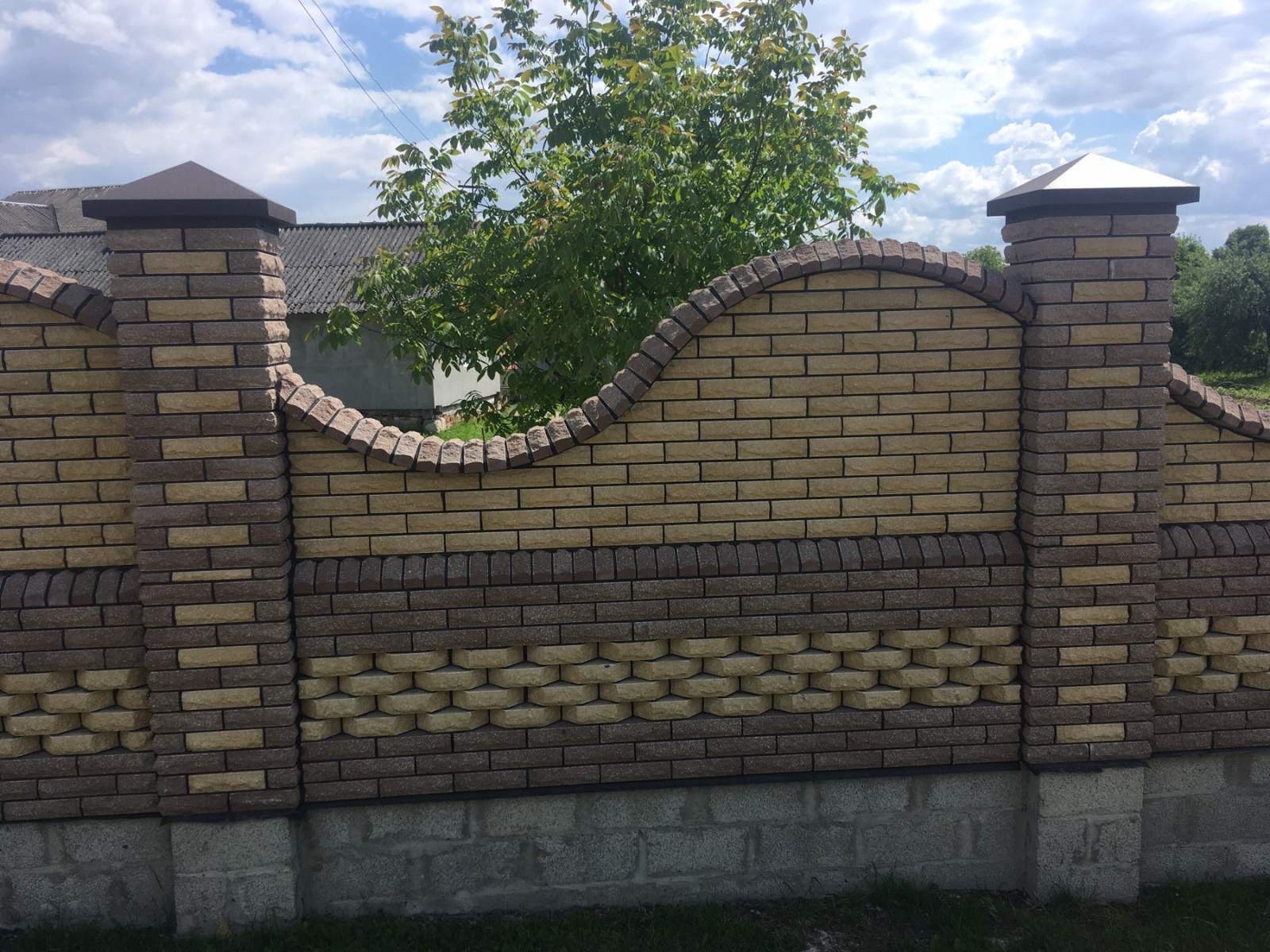 Dekoracyjny, murowany płot z cegły klinkierowej w odcieniach beżu i brązu, z ozdobnym łukiem i filarami zwieńczonymi daszkami. Widoczne elementy fundamentu.