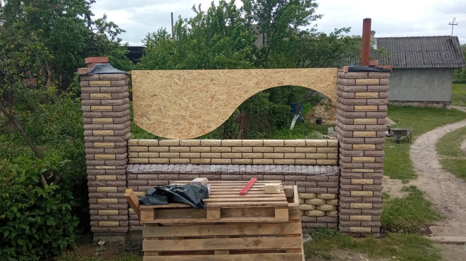 Wznoszona z cegły klinkierowej konstrukcja ogrodzenia z dekoracyjnym wycięciem w płycie OSB. Widoczne narzędzia i materiały budowlane na palecie. Tło stanowi zieleń i zabudowa.