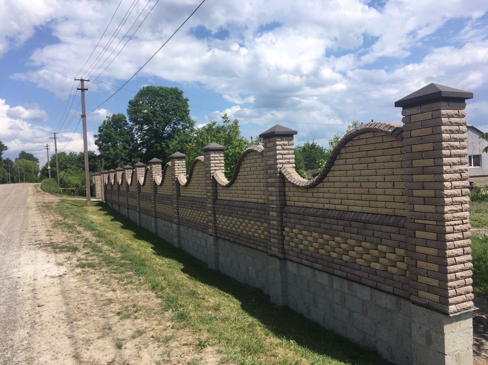 Falisty, dwukolorowy mur z cegły klinkierowej na betonowej podmurówce, zwieńczony daszkami. Widok na wiejski krajobraz z polną drogą i liniami energetycznymi.