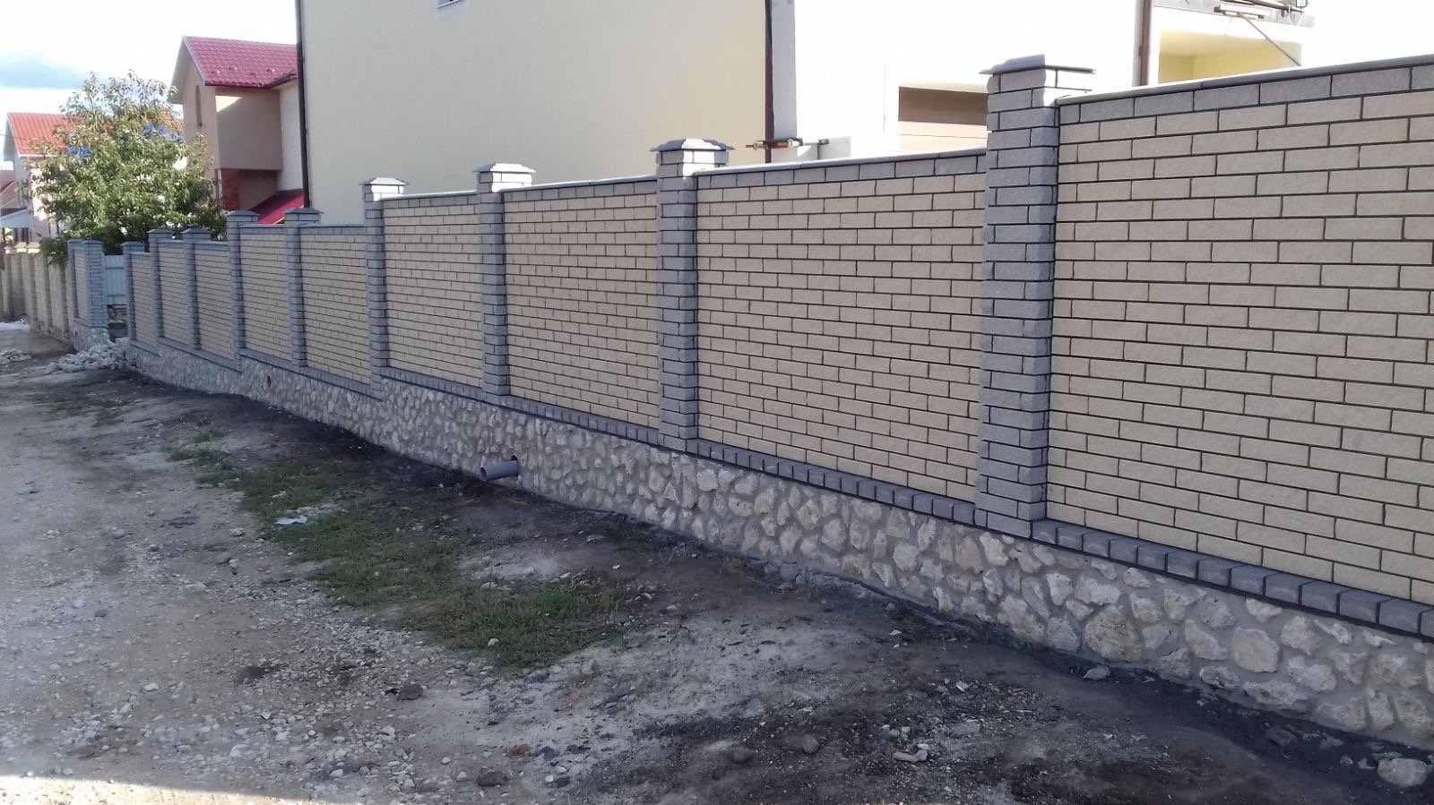 Nowoczesne ogrodzenie z jasnej cegły klinkierowej z szarymi słupkami i kamienną podmurówką, widoczne z poziomu gruntu. Równy teren, dom w tle.