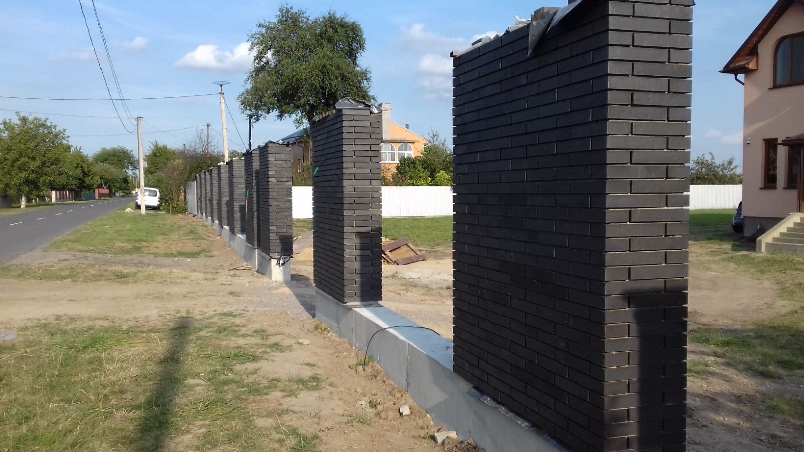 Ciemna, murowana podmurówka ogrodzenia z cegły klinkierowej, z widocznymi słupkami i fragmentami przęseł, na tle zieleni i zabudowań.