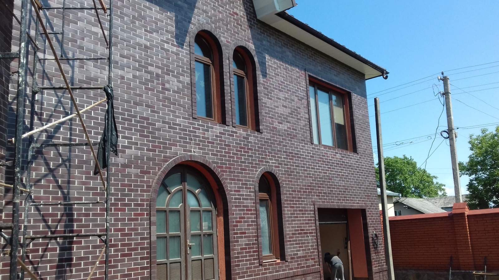 Dwupiętrowy dom z ciemnej cegły klinkierowej z łukowymi oknami, częściowo zasłonięty rusztowaniem. Widoczny fragment muru z cegły w tle i słup energetyczny.
