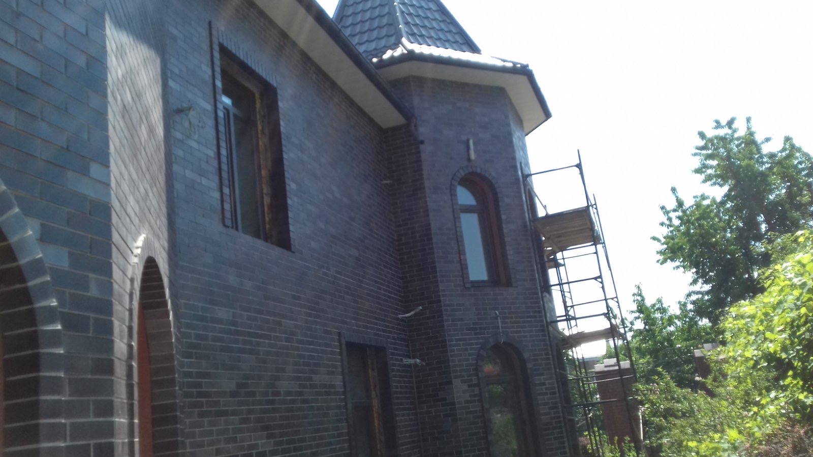 Elewacja z ciemnej cegły klinkierowej, widok z dołu na narożnik budynku z wieżyczką. Widoczne rusztowania i zieleń w tle. Charakterystyczne łukowe wnęki okienne.
