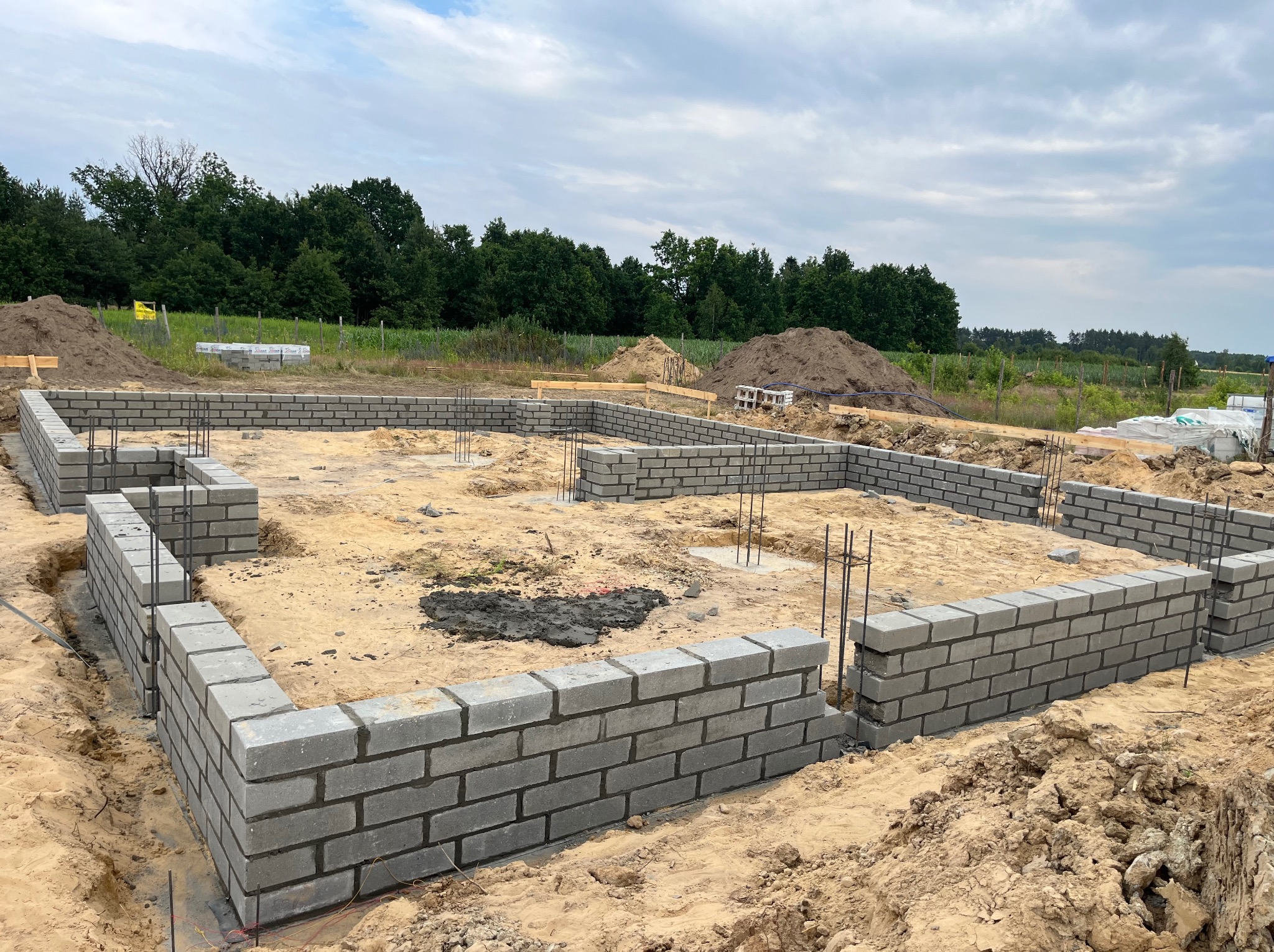 Fundamenty domu w trakcie budowy: ściany z szarych bloczków betonowych, wystające pręty zbrojeniowe, piasek i ziemia. W tle drzewa i pochmurne niebo.