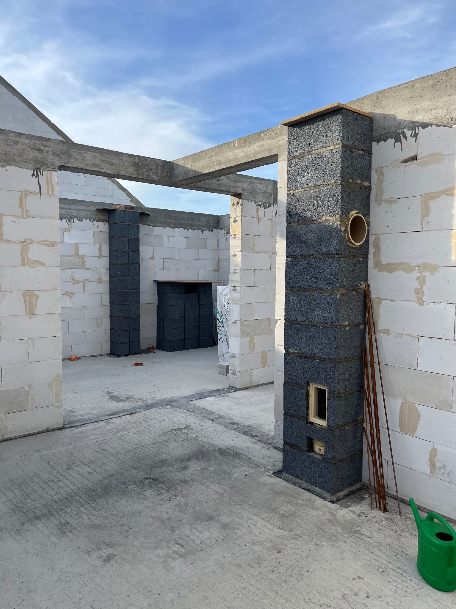 Wnętrze surowego domu w trakcie budowy: ściany z białych bloczków, betonowe nadproża, komin z szarych bloczków, pręty zbrojeniowe i zielona konewka.