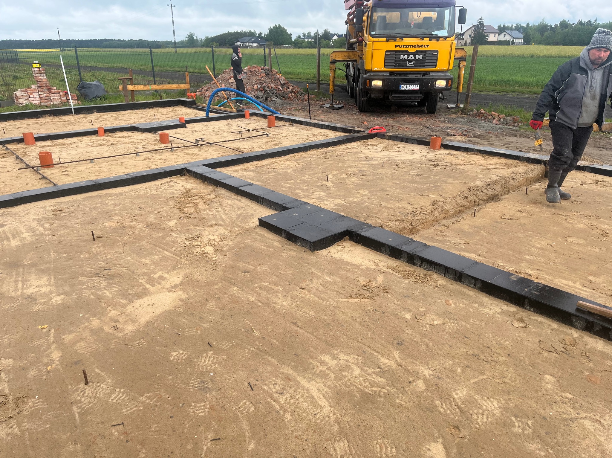 Przygotowany teren pod fundamenty z ułożonymi bloczkami, widoczne rury kanalizacyjne i zbrojenie. W tle betoniarka i pracownik budowlany.