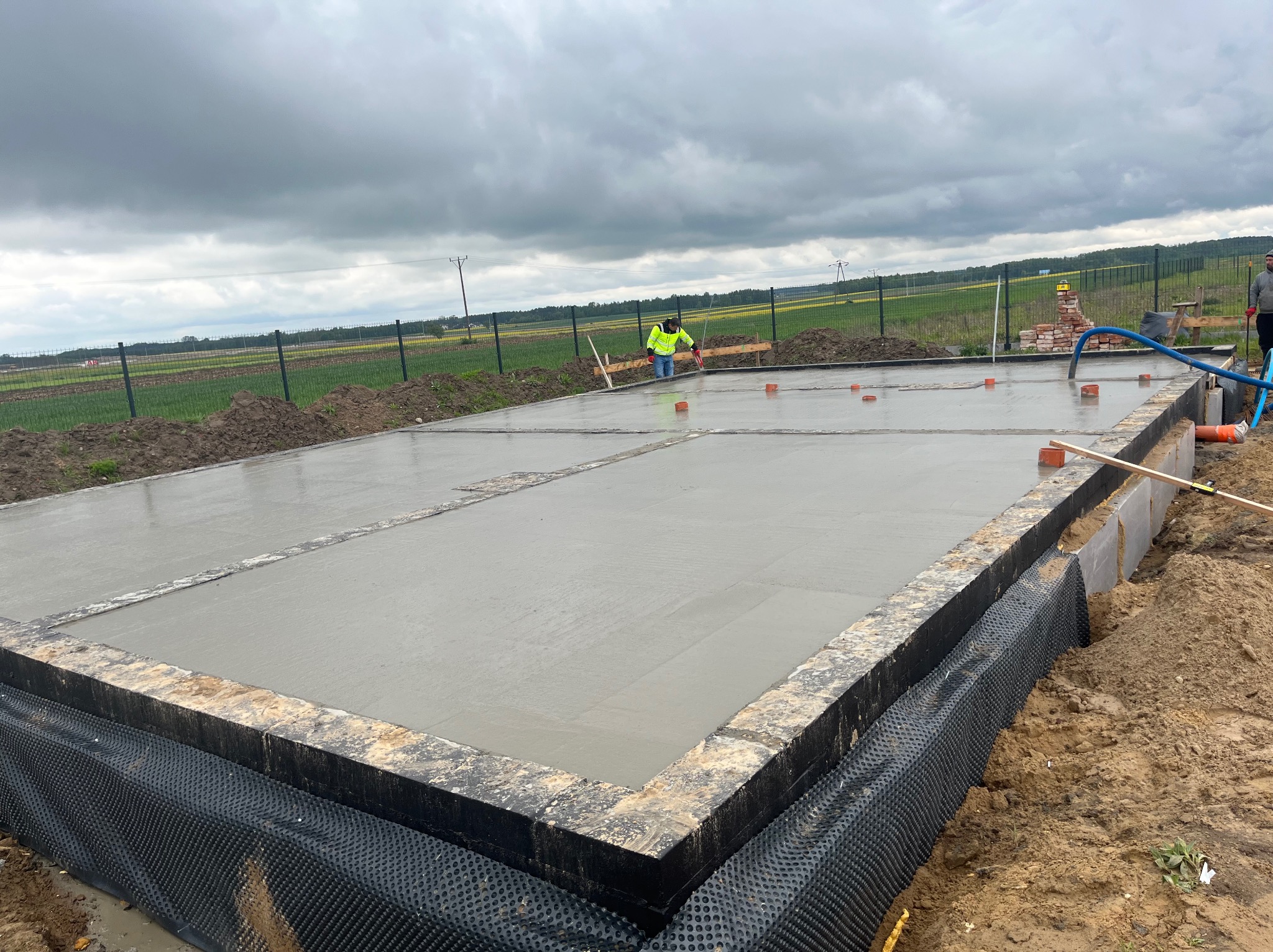 Zalany fundament z izolacją pionową, widoczny pracownik wyrównujący powierzchnię betonu. W tle krajobraz polny pod pochmurnym niebem.