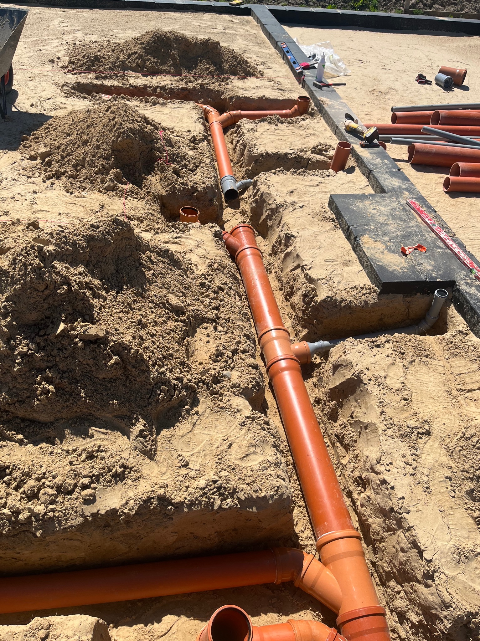 Pomarańczowe rury kanalizacyjne ułożone w wykopie z piasku, widoczne narzędzia i poziomica. Etap prac instalacyjnych przed zasypaniem.