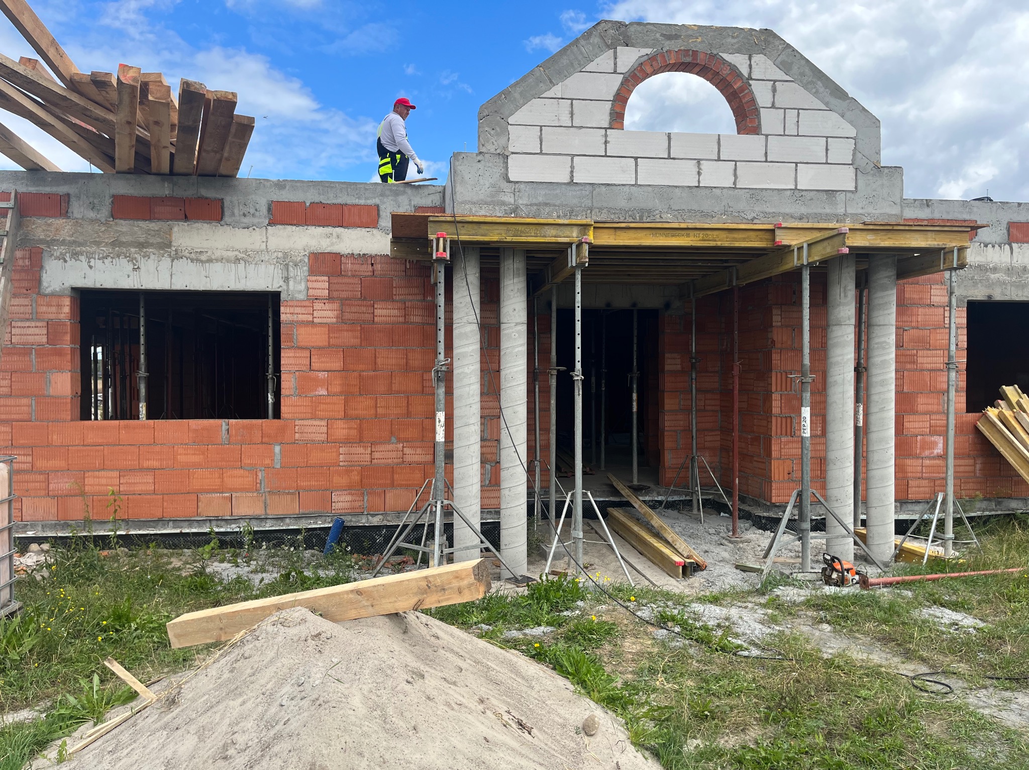 Budowa domu z czerwonej cegły, widoczne betonowe kolumny, szalunki, piasek i pracownik na dachu. W tle błękitne niebo z chmurami.