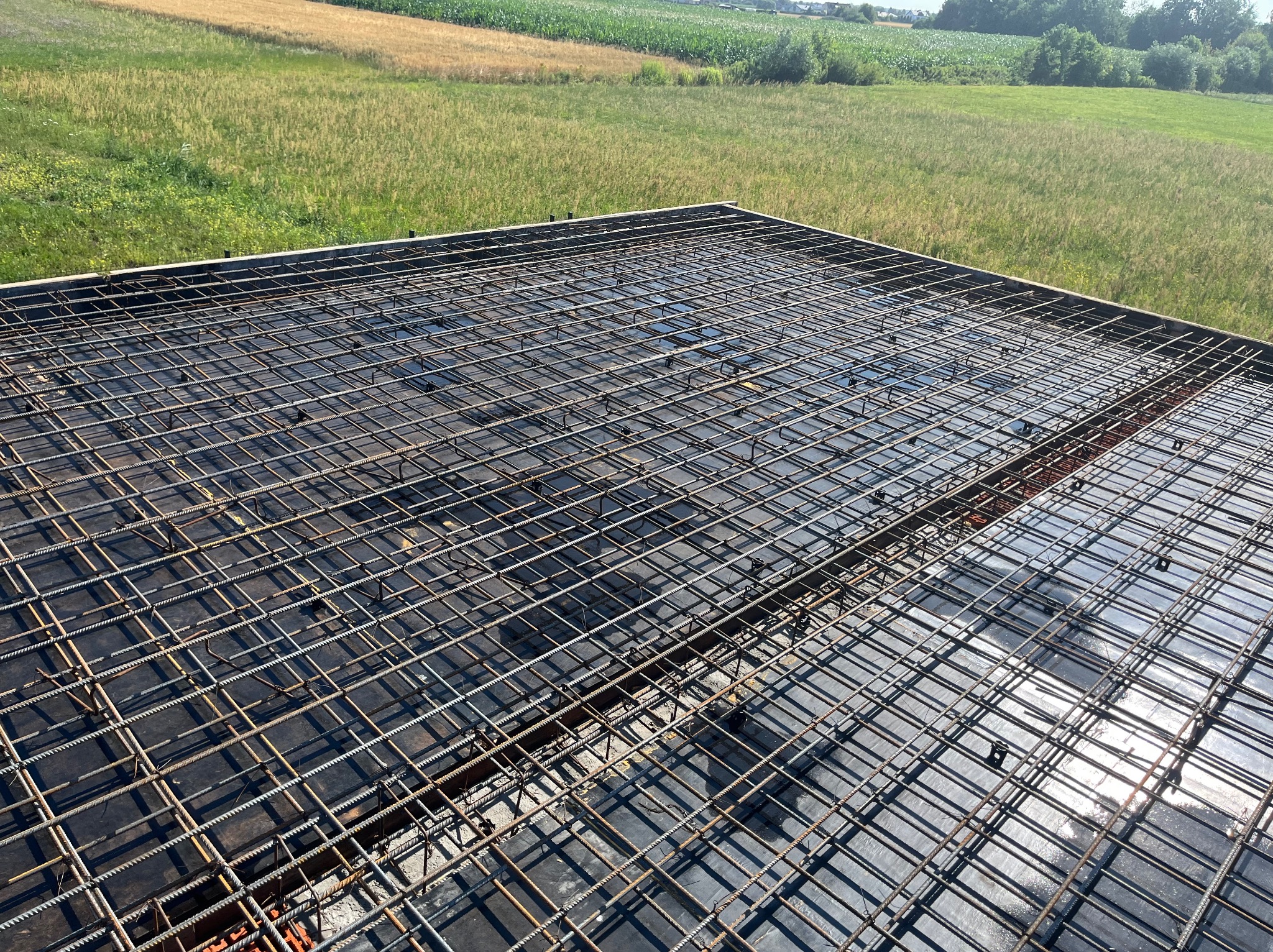 Zbrojenie stropu na czarnej folii, przygotowane do zalania betonem. W tle zielone pole i drzewa. Widok z góry, oświetlenie słoneczne.