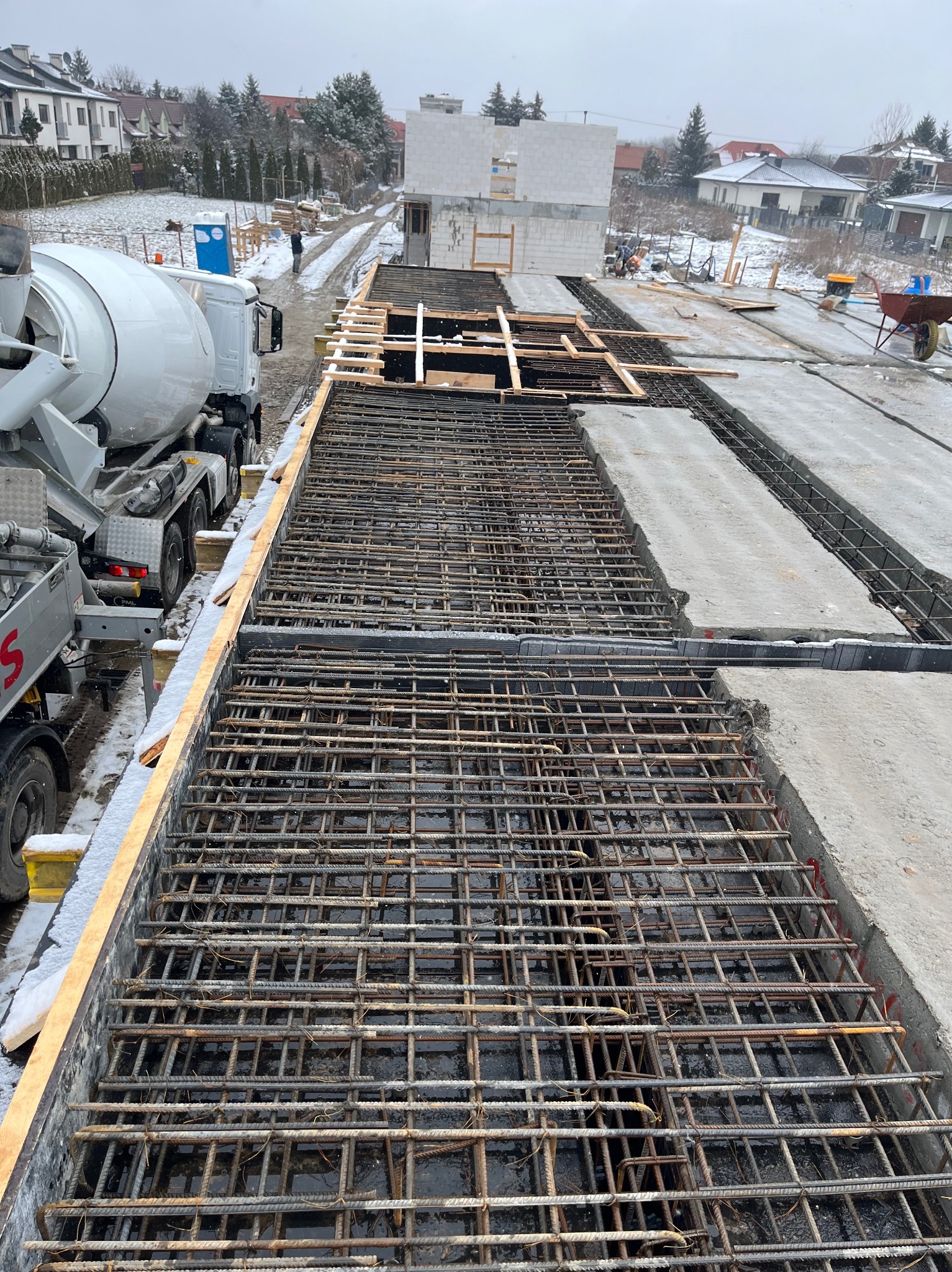 Zbrojenie fundamentów pod budowę domu jednorodzinnego, widok z góry. Na pierwszym planie siatka zbrojeniowa, w tle beton i betoniarka. Zimowa sceneria.