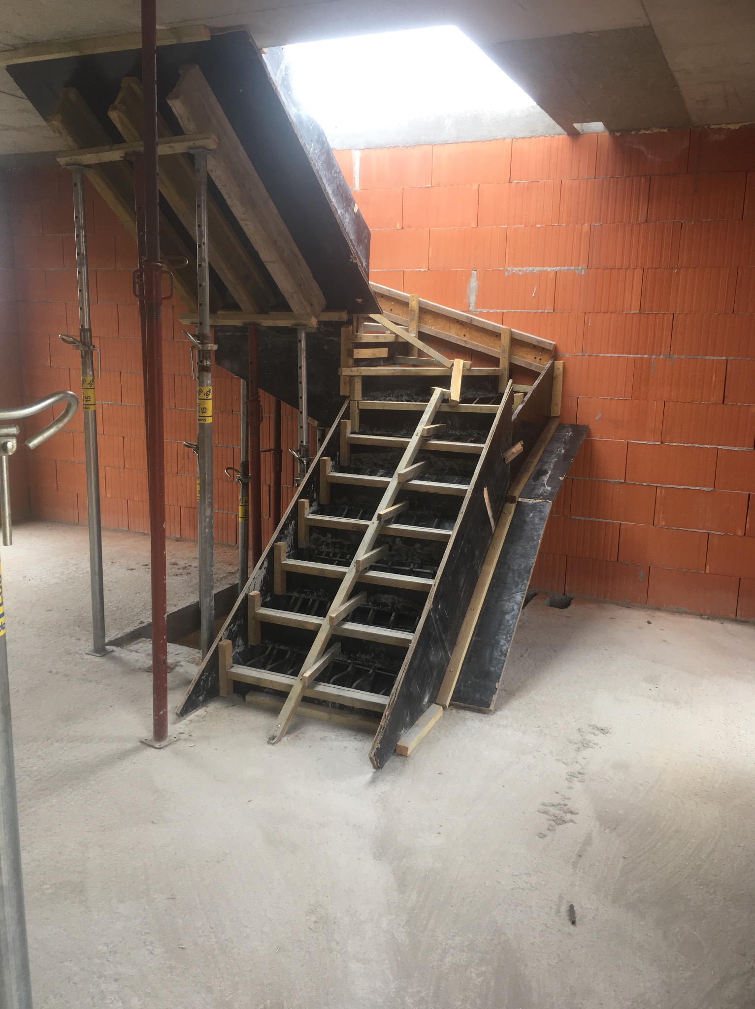 Szare, betonowe schody w trakcie budowy, z drewnianym szalunkiem i stalowym zbrojeniem, na tle pomarańczowej ściany z pustaków. Widoczne podpory i fragment sufitu z oknem.