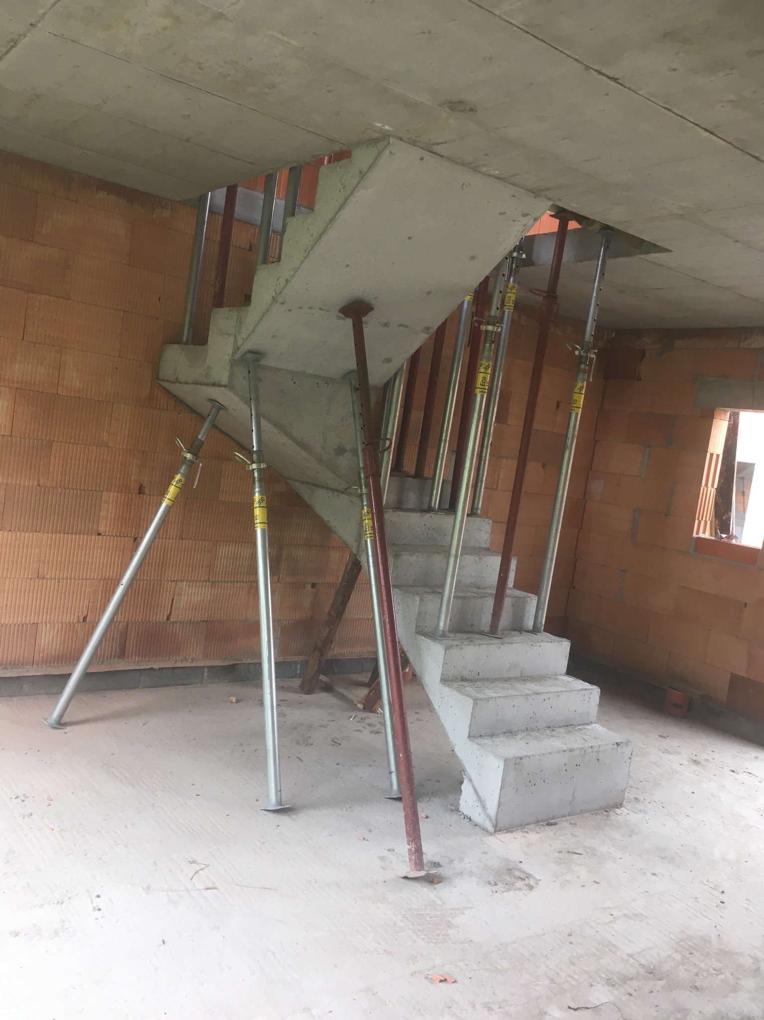 Betonowe schody w surowym stanie, podparte metalowymi stemplami wewnątrz budynku z czerwonej cegły. Widoczne otwory w stropie i fragment okna.