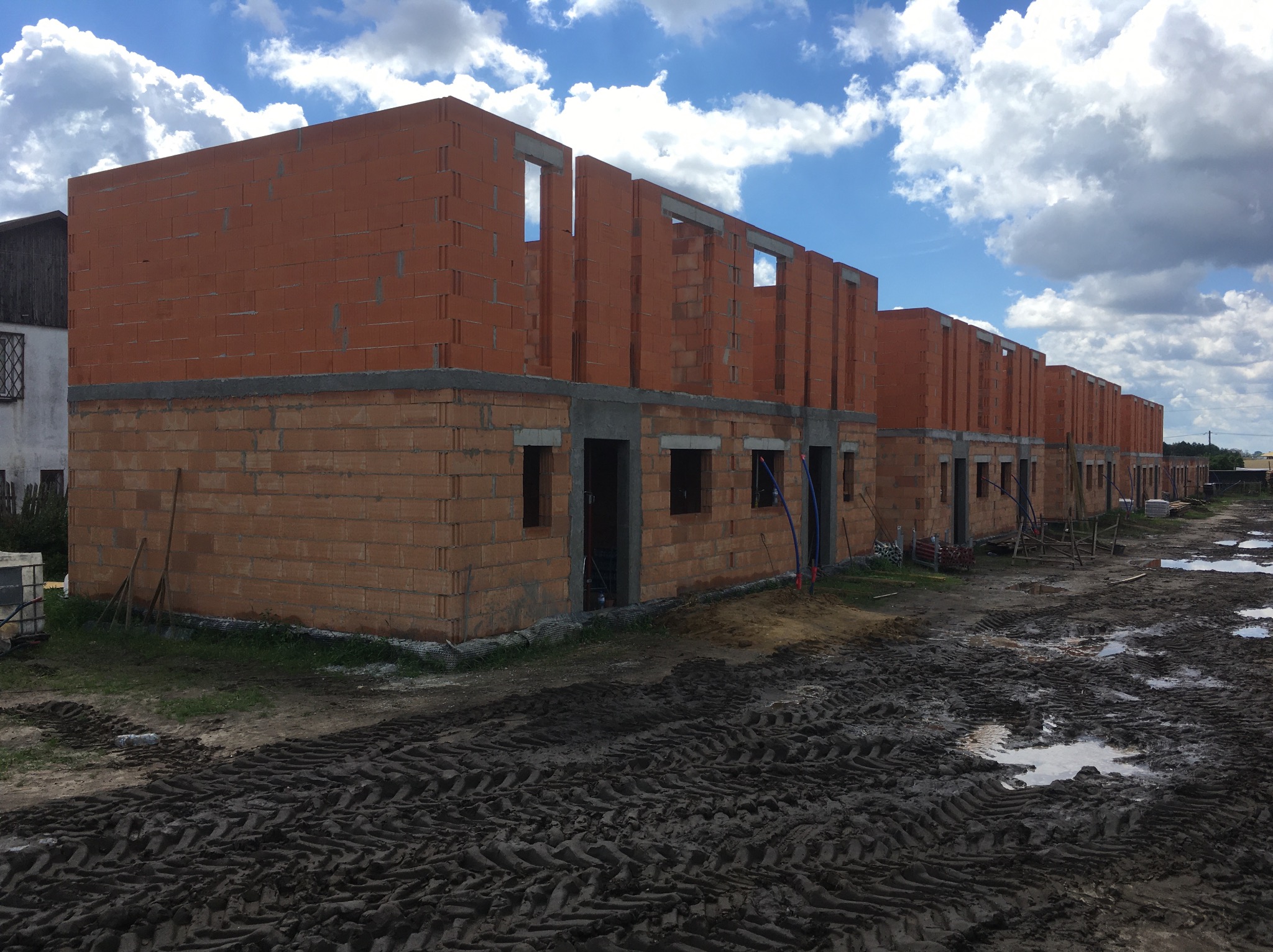 Rząd surowych, ceglanych budynków w trakcie budowy pod błękitnym niebem z chmurami. Widoczne ślady opon na błotnistym terenie.