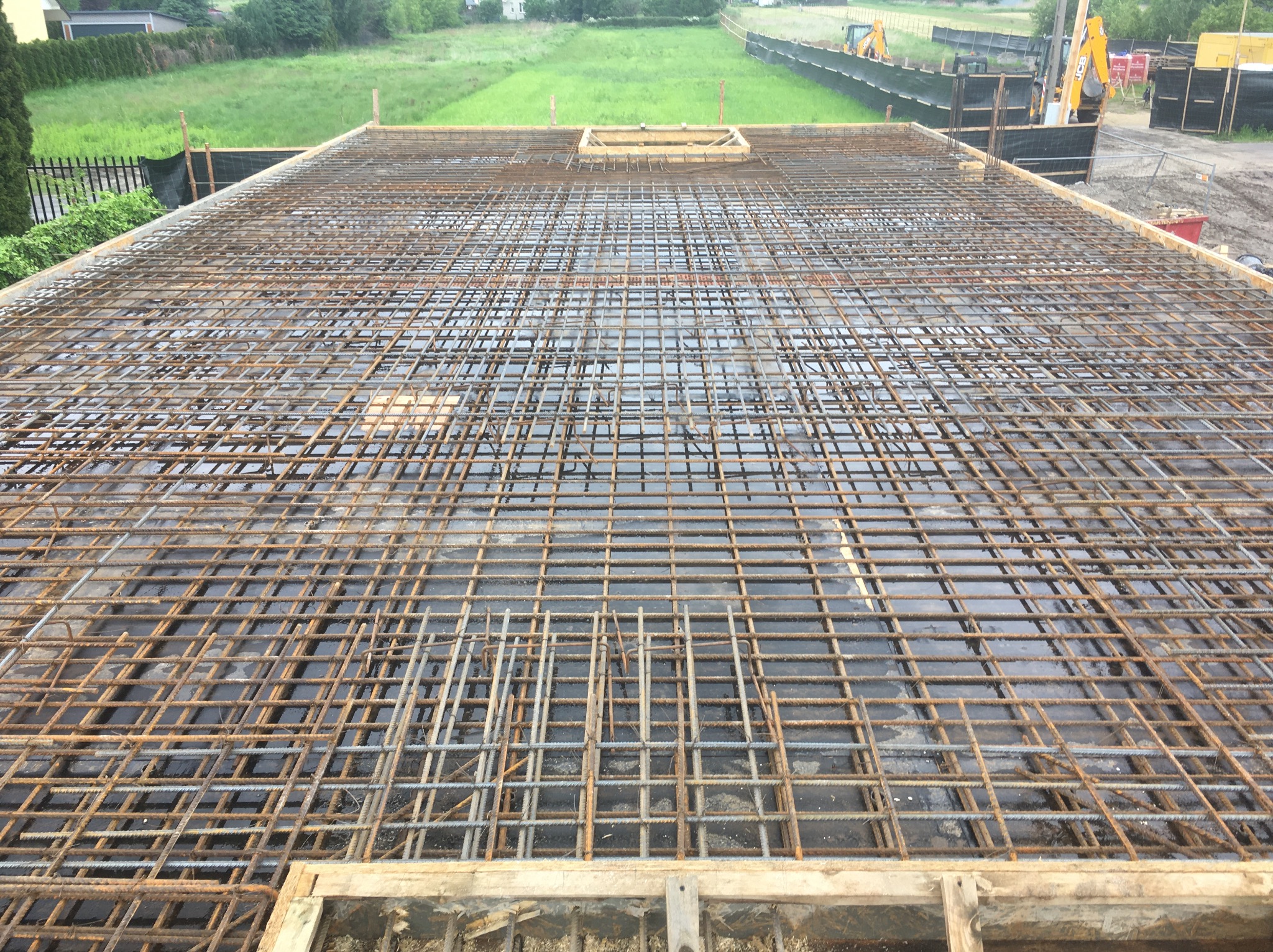 Zbrojenie płyty fundamentowej z widocznym deskowaniem i otoczeniem budowy. Widok z góry na siatkę z prętów zbrojeniowych, przygotowaną do zalania betonem.