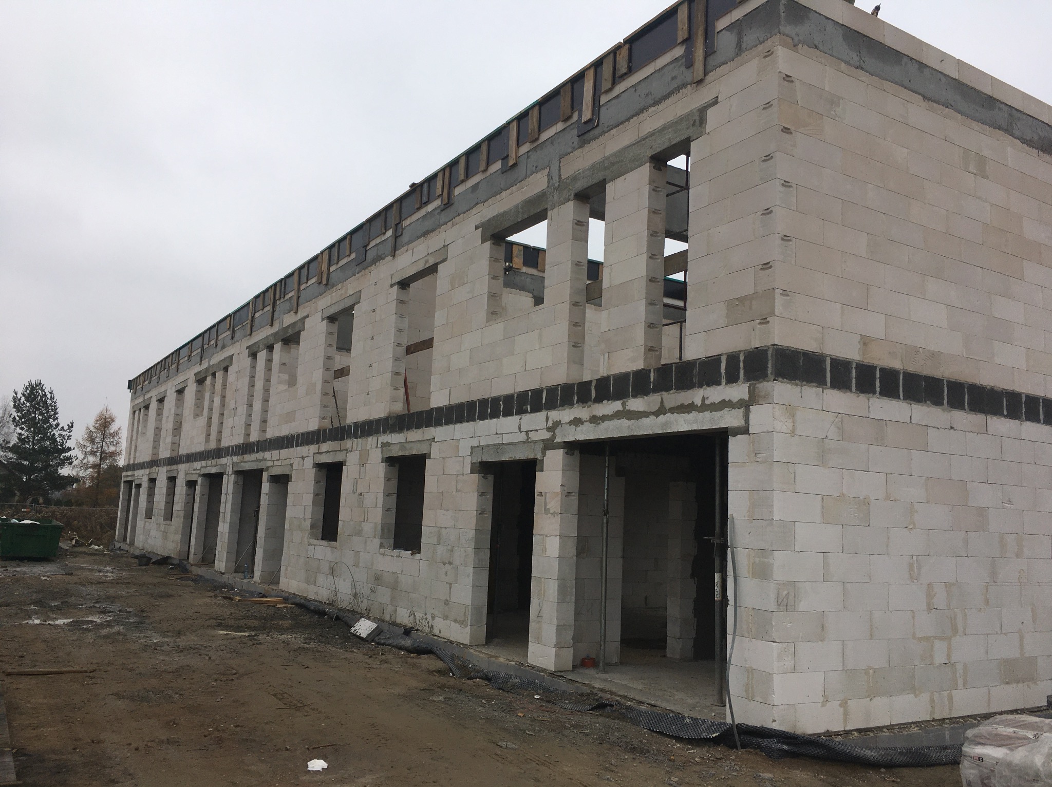 Wznoszenie ścian z betonu komórkowego na budowie wielorodzinnego budynku. Widoczne otwory okienne i drzwiowe, szalunki stropowe. Szary dzień.