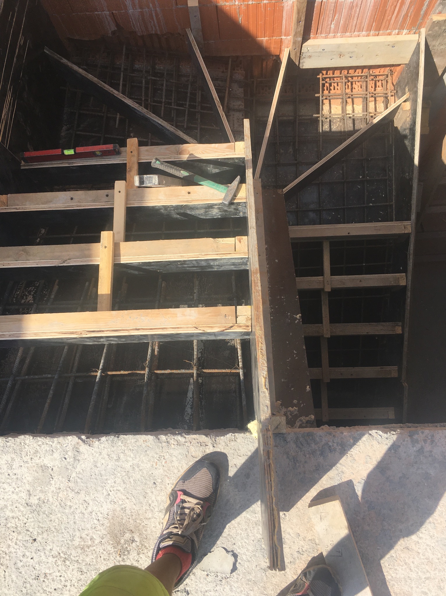 Zbrojenie fundamentów w szalunku z desek, widok z góry. Poziomica, młotek i buty robocze w kadrze. Elementy konstrukcyjne z betonu i cegły.