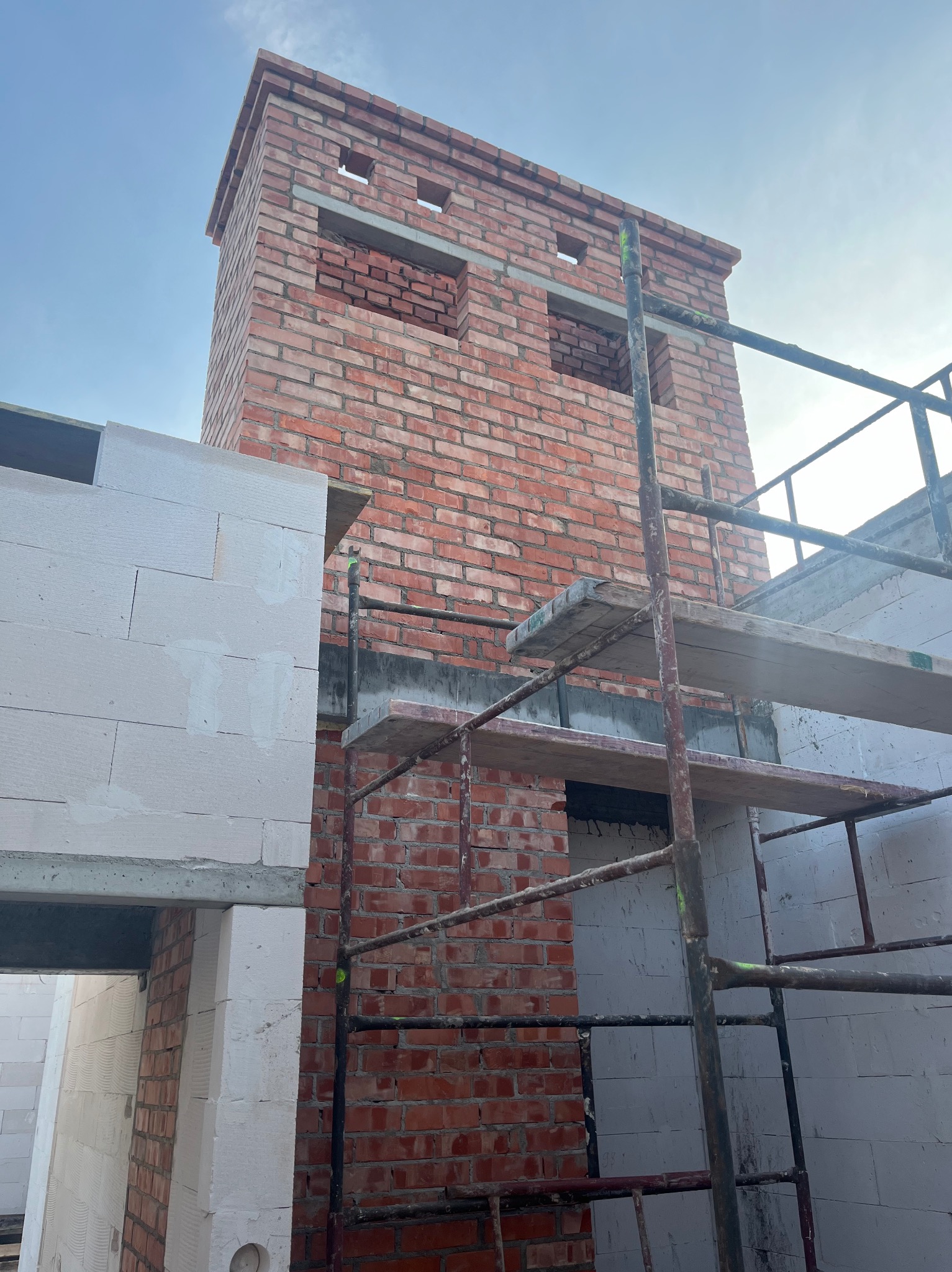 Wznoszenie komina z czerwonej cegły na tle błękitnego nieba, widoczne rusztowanie i fragmenty ścian z białego betonu komórkowego. Etap budowy w toku.