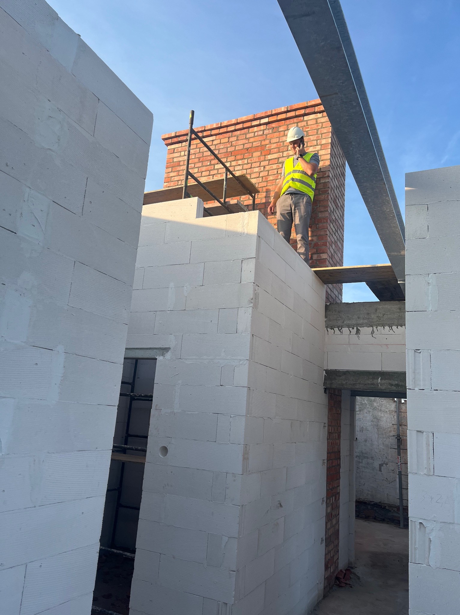Wznoszenie murów z białych bloczków i ceglanego komina. Pracownik w kasku i kamizelce na tle błękitnego nieba. Widoczne rusztowania i stalowe belki stropowe.