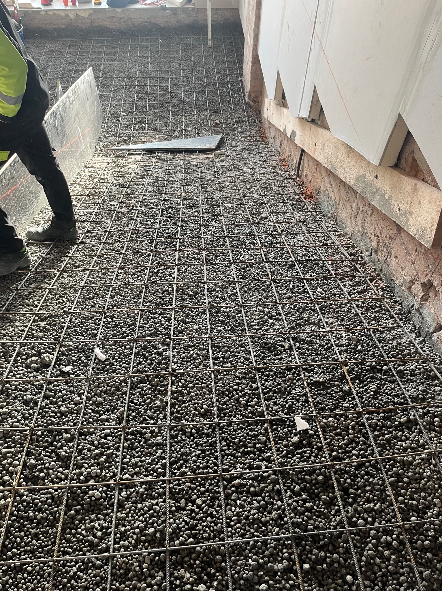 Przygotowanie podłoża z izolacją termiczną i siatką zbrojeniową przed wylewką. Widoczne płyty izolacyjne przy ścianie i fragment pracownika w odblaskowej kamizelce.