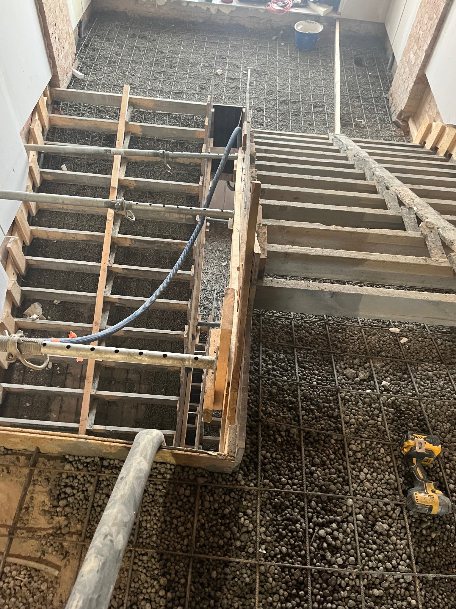 Z góry widok na szalunek zbrojony siatką i wypełniony kruszywem, z widocznym wężem do betonu i wiertarką akumulatorową. Etap prac budowlanych.