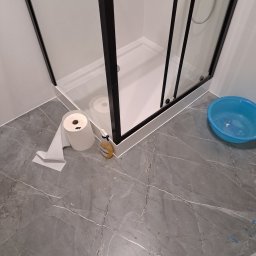 Tomasz Szydlik Usługi Remontowo-budowlane - Nowoczesna kabina prysznicowa z czarnymi profilami na tle szarych płytek imitujących marmur. Widoczne akcesoria łazienkowe i niebieska miska.