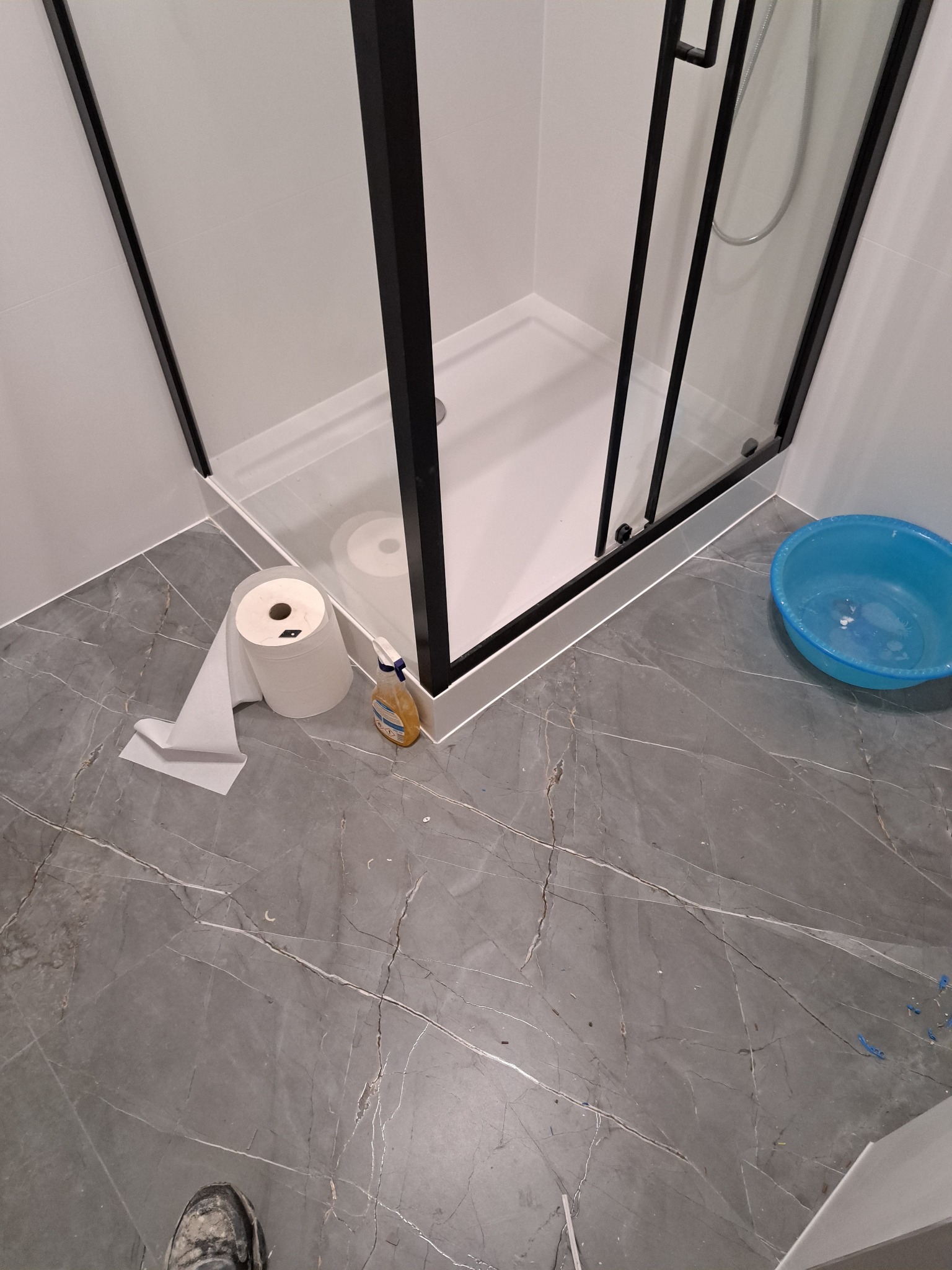 Nowoczesna kabina prysznicowa z czarnymi profilami na tle szarych płytek imitujących marmur. Widoczne akcesoria łazienkowe i niebieska miska.