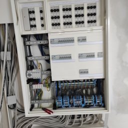 Firma Usługowa Raupe Daniel Gąsienica - Otwarta, biała skrzynka rozdzielcza z licznymi bezpiecznikami, przewodami i elementami montażowymi. Widoczne narzędzia, kable w ścianie. Ujęcie z bliska, w trakcie montażu.