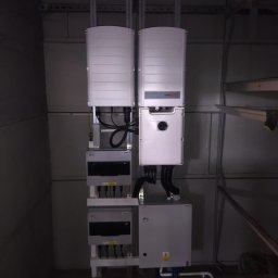 Kompletna instalacja fotowoltaiczna w pomieszczeniu technicznym. Widoczne falowniki, skrzynki przyłączeniowe i okablowanie. Elementy zamontowane na ścianie w szarym pomieszczeniu.