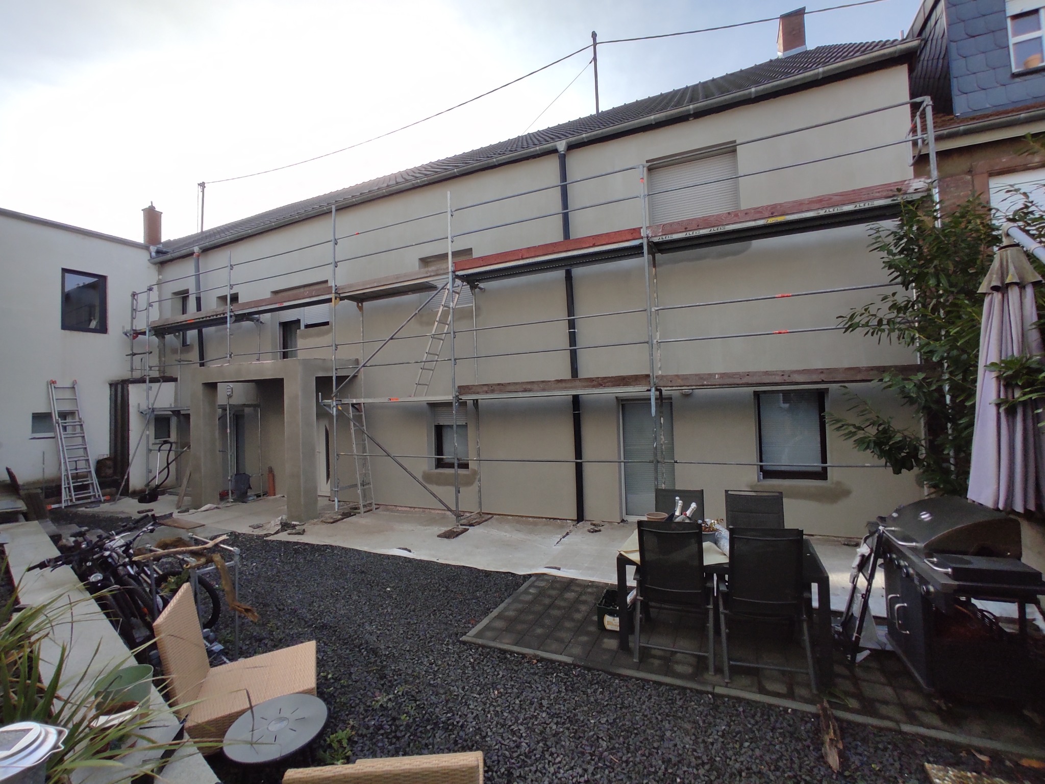 Elewacja budynku w trakcie prac wykończeniowych, widoczne rusztowania i świeżo nałożony tynk. W tle elementy wyposażenia ogrodu, w tym meble i grill.