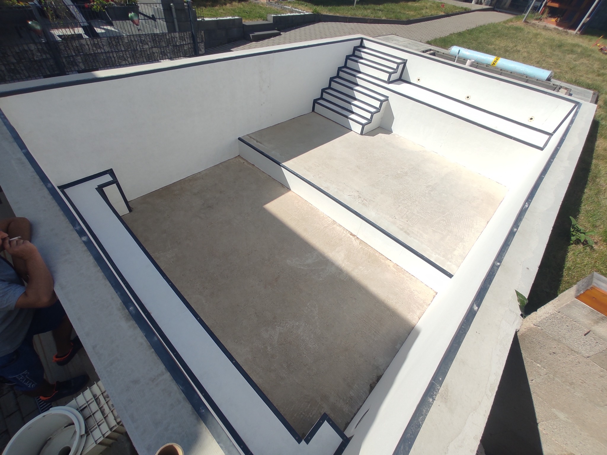 Nowoczesny, betonowy basen w trakcie budowy, z widocznymi schodami i wykończeniami w kolorze białym i czarnym. Widok z góry, oświetlenie słoneczne.