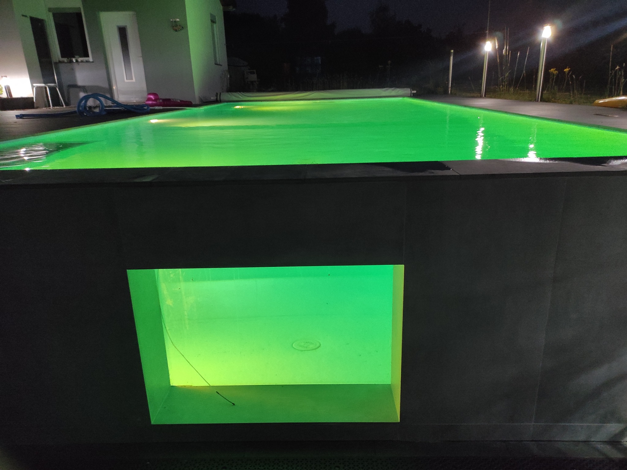 Nocny widok na basen z podświetleniem LED, woda oświetlona na zielono, widoczne lampy oświetlające teren wokół basenu, nowoczesny design.