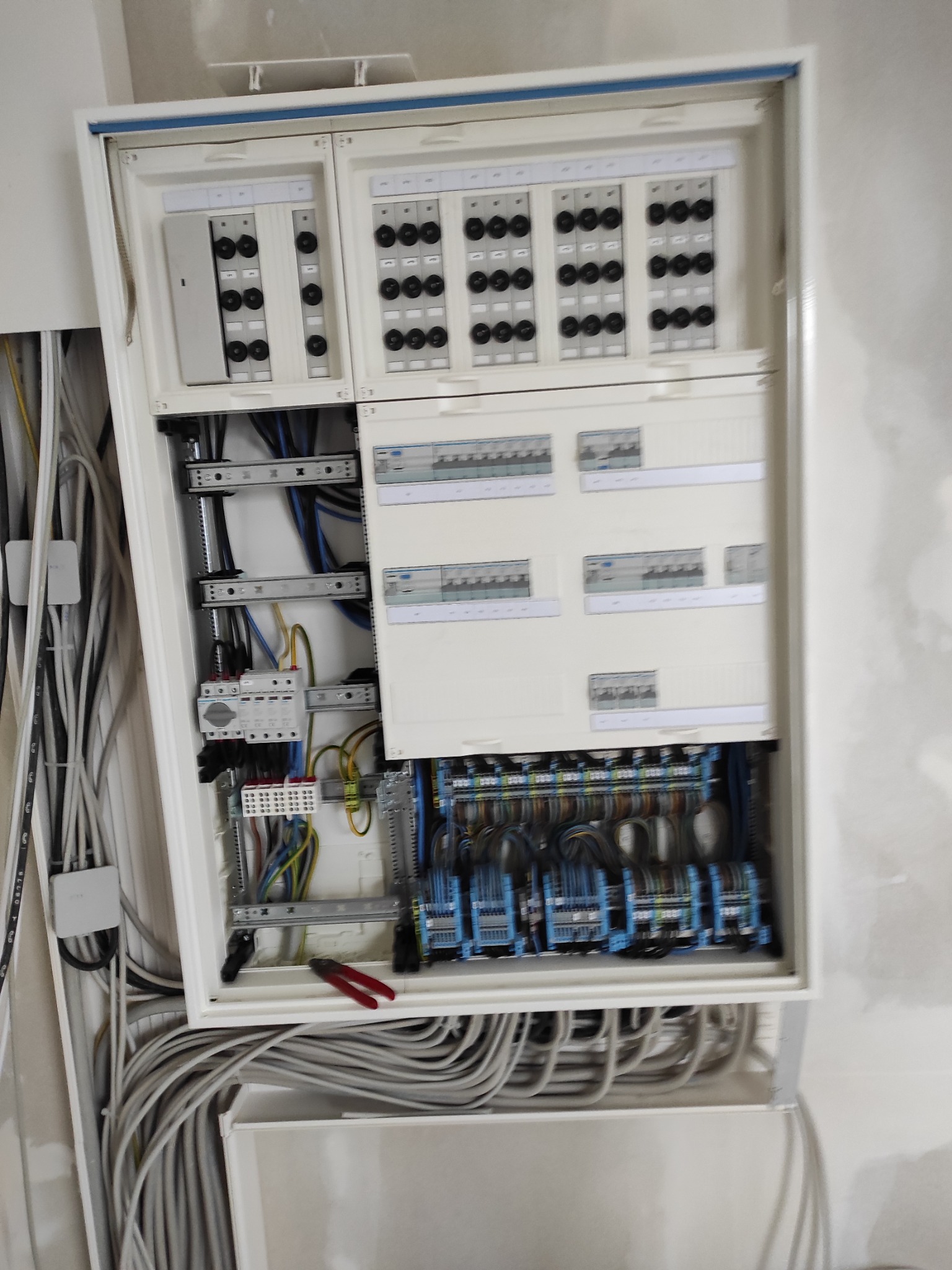 Otwarta, biała skrzynka rozdzielcza z licznymi bezpiecznikami, przewodami i elementami montażowymi. Widoczne narzędzia, kable w ścianie. Ujęcie z bliska, w trakcie montażu.