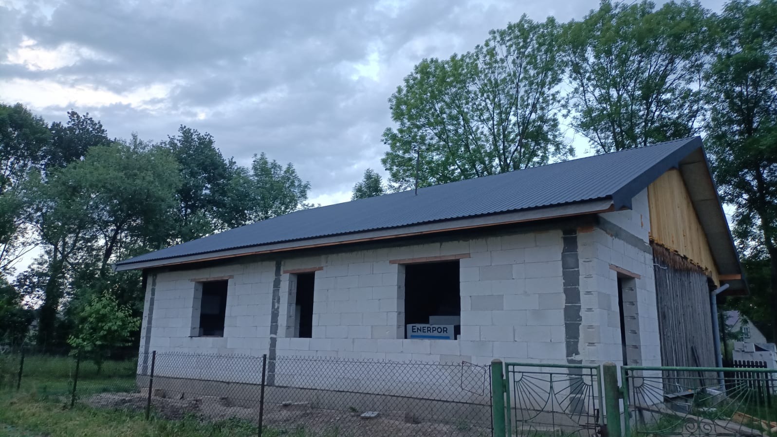 Dom w trakcie budowy z szarego bloczka, z zamontowanym, ciemnym dachem z blachy falistej. Widoczne niezabudowane okna i drzwi. Tło stanowią drzewa i pochmurne niebo.