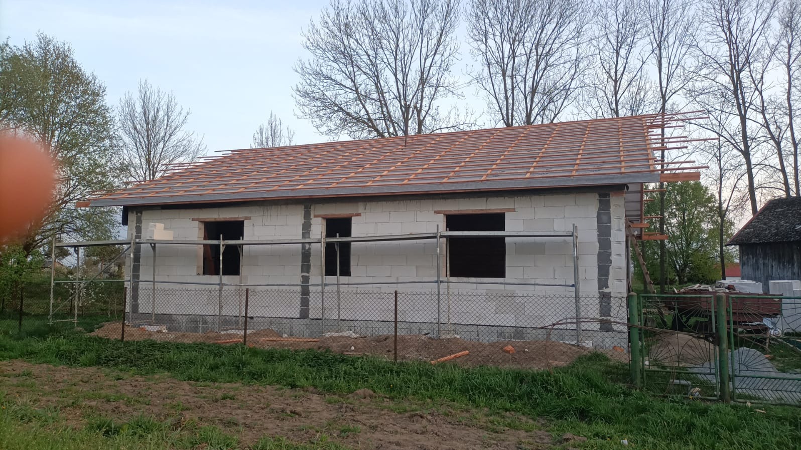 Budowa domu jednorodzinnego: ściany z betonu komórkowego, więźba dachowa w trakcie montażu, widoczne rusztowania i teren budowy. Ujęcie z zewnątrz.