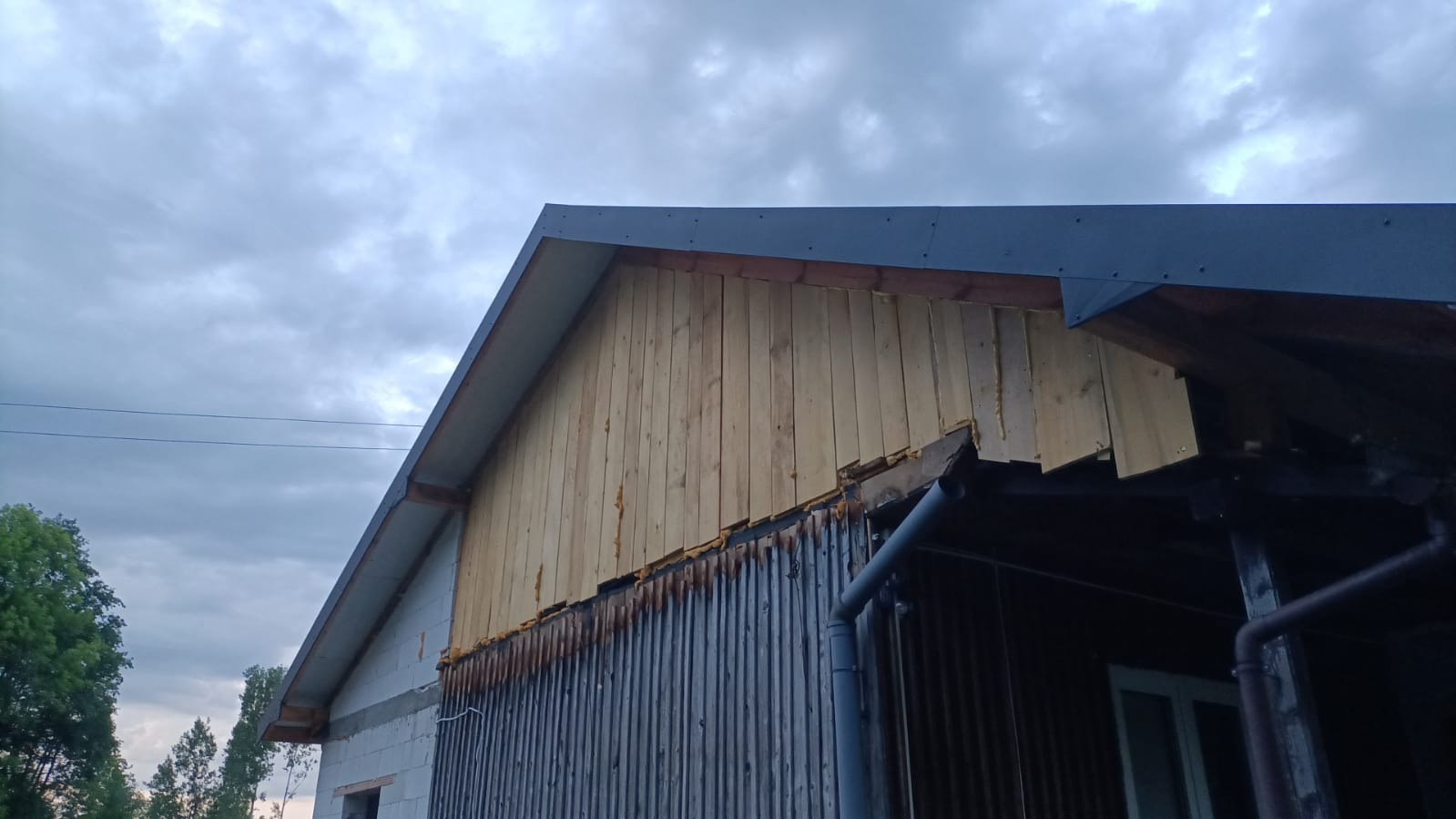 Fragment elewacji domu w trakcie prac: drewniane deski na szczycie ściany, widoczne elementy konstrukcyjne dachu i rynna. Częściowo obita ściana budynku.