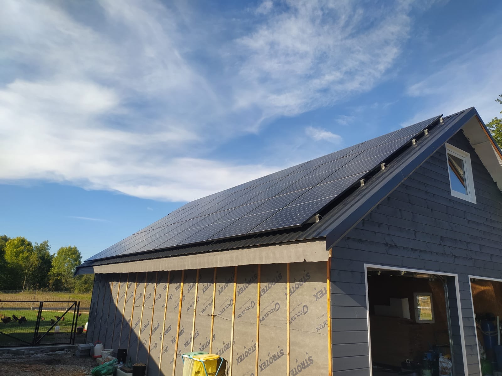 Montaż paneli fotowoltaicznych na dachu budynku z szarą elewacją. Widoczne membrany dachowe i elementy konstrukcyjne. Błękitne niebo z chmurami w tle.