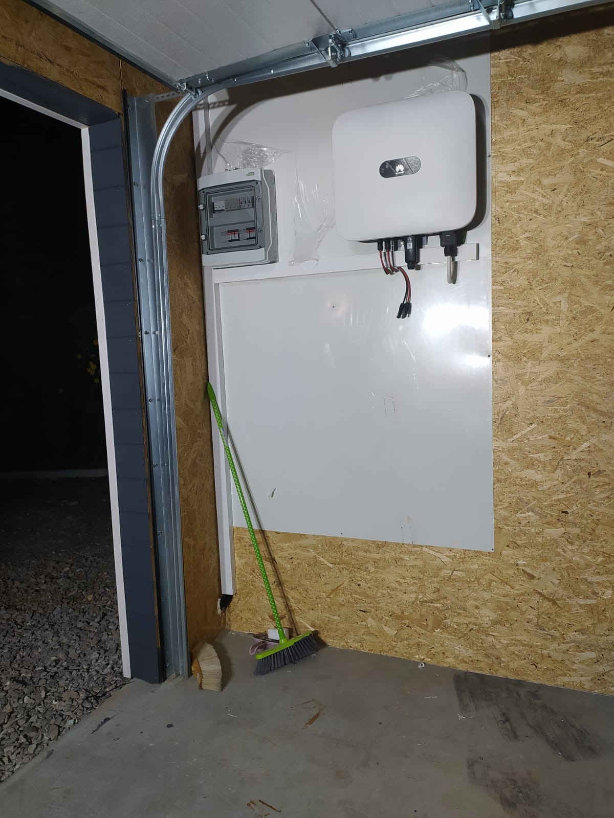 Wnętrze garażu z zamontowanym inwerterem fotowoltaicznym Huawei i skrzynką elektryczną na ścianie z płyty OSB. Obok ściany stoi zielona zmiotka.