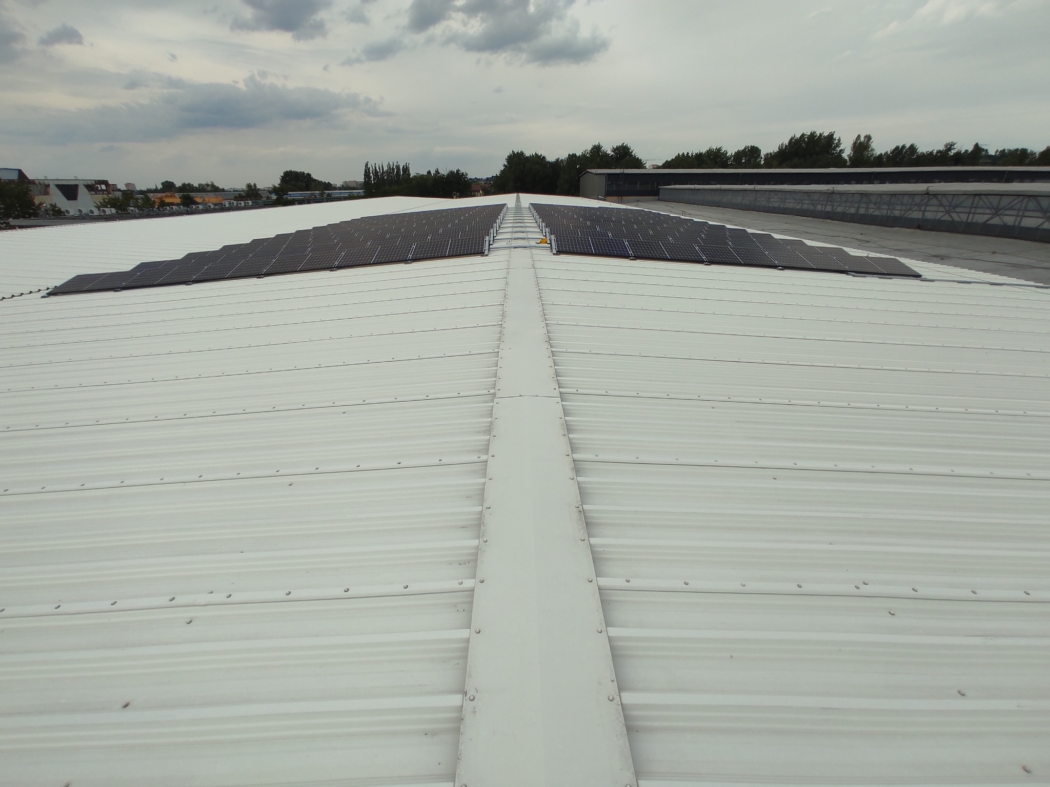 Biały, blaszany dach z zamontowanymi panelami fotowoltaicznymi w dwóch rzędach. Widok z góry, pochmurne niebo w tle. Metalowe łączenia na dachu.