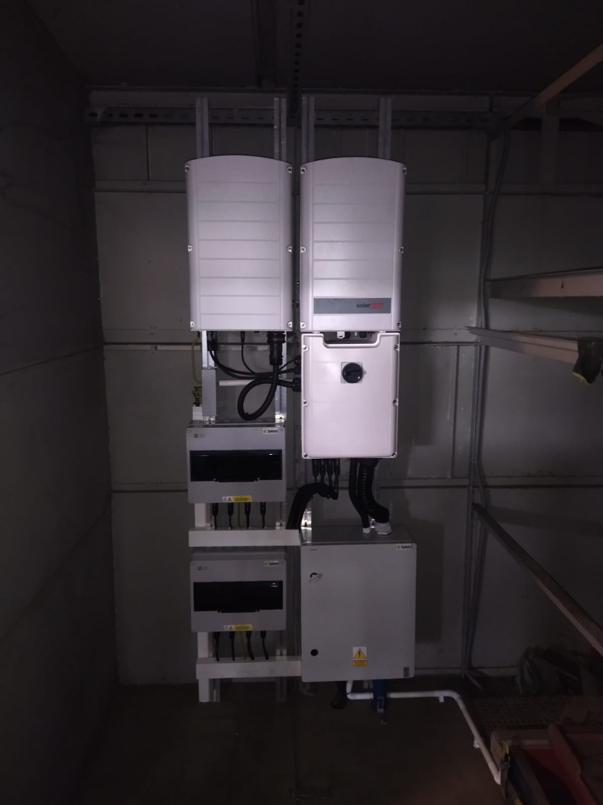 Kompletna instalacja fotowoltaiczna w pomieszczeniu technicznym. Widoczne falowniki, skrzynki przyłączeniowe i okablowanie. Elementy zamontowane na ścianie w szarym pomieszczeniu.