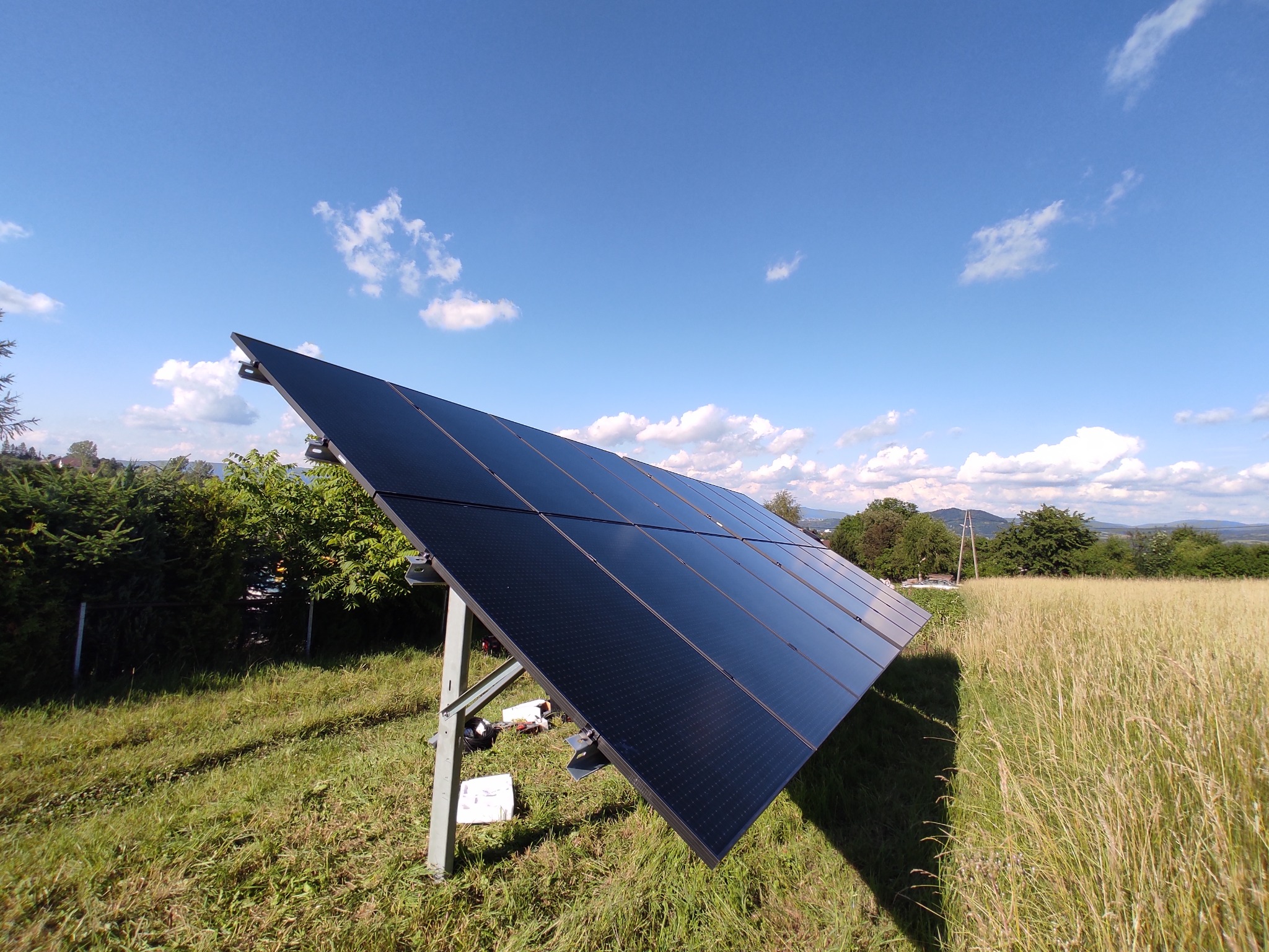 Panel fotowoltaiczny na łące w słoneczny dzień. Widoczne mocowania i elementy instalacji. Trawa wysoka, w tle drzewa i błękitne niebo z chmurami.