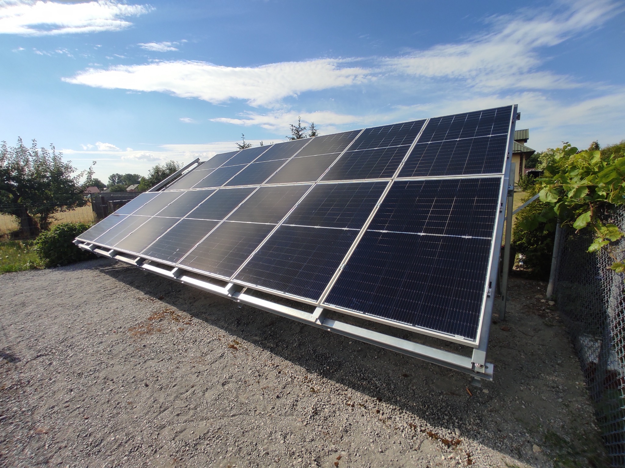 Panel fotowoltaiczny na metalowej konstrukcji, ustawiony pod kątem na żwirowym podłożu. W tle błękitne niebo z chmurami i zieleń drzew.